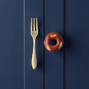 Puede incluir: Un tirador de armario de madera clara con forma de tenedor colocado verticalmente junto a un pomo de armario redondo y marrón con forma de dónut. Ambas piezas están montadas en una puerta de armario azul marino con paneles verticales.