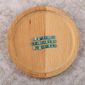 Puede incluir: Una bandeja redonda de madera con acabado marrón claro. Tres filas de pequeñas piedras preciosas cuadradas de color turquesa están dispuestas en la bandeja. Las piedras preciosas tienen una forma ligeramente irregular y una superficie pulida.