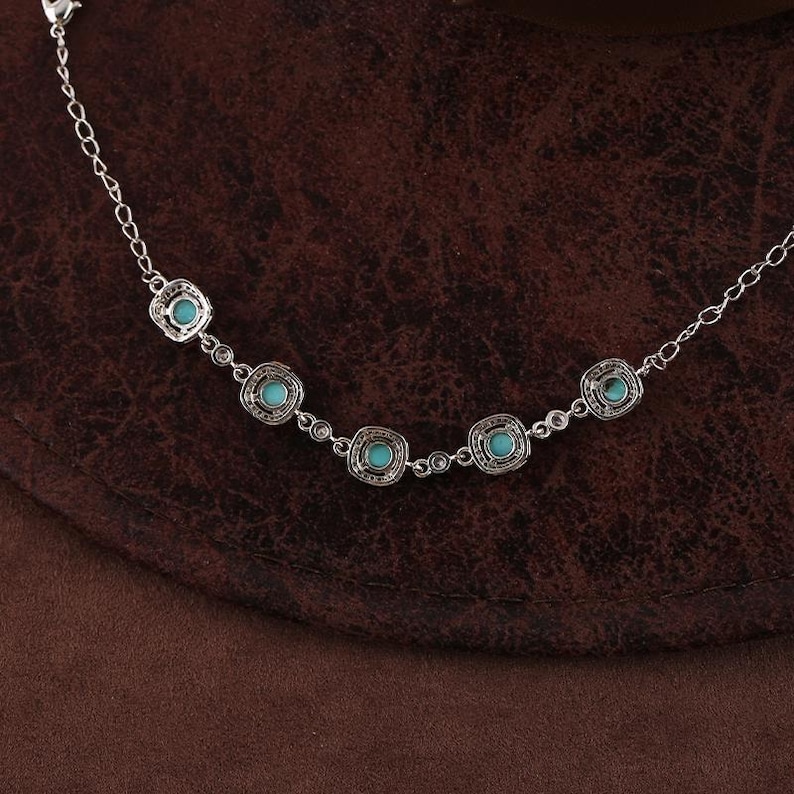 Pode incluir: Uma pulseira prateada com cinco pedras quadradas de cor turquesa. A pulseira tem uma corrente delicada e est&aacute; colocada sobre um fundo castanho. As pedras est&atilde;o engastadas numa moldura decorativa vazada.