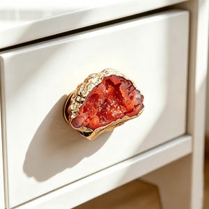 May include: A decorative drawer pull with a gold-colored, textured frame and a vibrant red, crystal-like center. The pull is mounted on a white drawer, creating a striking contrast. The design resembles a geode.