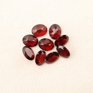 Può includere: Una collezione di otto pietre preziose rosso scuro, di forma ovale e sfaccettate, disposte in un gruppo circolare. Ogni pietra presenta una superficie lucida con molteplici sfaccettature geometriche che riflettono la luce, mostrando un colore cremisi uniforme.