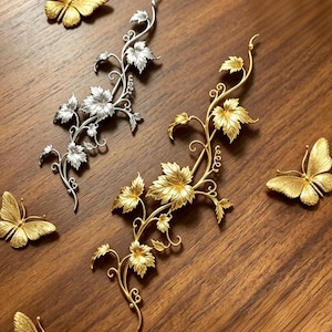 May include: Two metal vine-shaped decorative pieces, one silver-toned and one gold-toned, featuring detailed leaves and curling tendrils. They are surrounded by four gold-toned butterfly-shaped decorative pieces with textured wing patterns.
