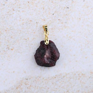 May include: A rough, dark red garnet pendant with a gold-coloured bail. The gemstone has a rough, natural texture and is suspended from a simple, elegant gold-tone loop. The pendant is a unique piece of jewellery.