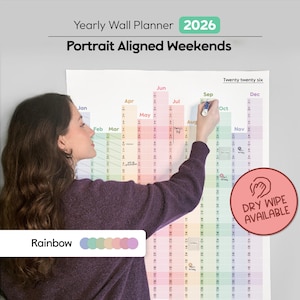 Peut inclure: Un grand planificateur mural annuel 2026 de couleur arc-en-ciel avec des week-ends alignés en portrait. Une personne écrit sur le planificateur avec un marqueur bleu. Le texte "Dry Wipe Available" est visible. Le planificateur a le texte "Yearly Wall Planner 2026".