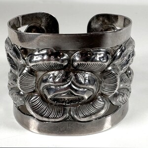 May include: A silver bracelet with a floral design. The bracelet is made of thick, textured silver and has a wide, open design.