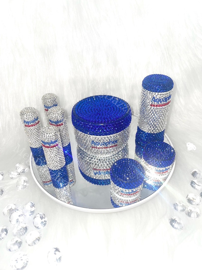 以下が含まれることがあります： ラインストーンが付いた青と銀の容器5個セット。容器は鏡面トレーの上に置かれています。容器には「Aquaphor」の文字が書かれています。