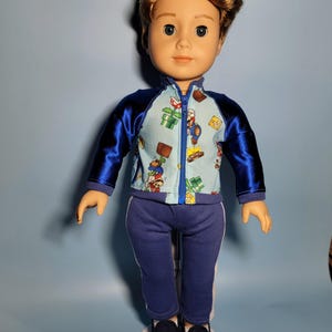 May include: A blue and white tracksuit with a Mario-themed print on the jacket. The jacket has a zipper and blue satin sleeves. The doll is wearing blue sneakers.