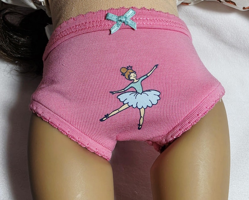May include: Pink underwear with a light blue bow and a ballerina graphic. The underwear has a scalloped edge and a cartoon ballerina in a blue tutu. The underwear is made of a soft, ribbed fabric.