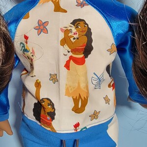 May include: A doll wearing a blue and white outfit with a character design. The top is white with a blue trim and features a cartoon illustration. The pants are solid blue. The outfit is designed for a doll.
