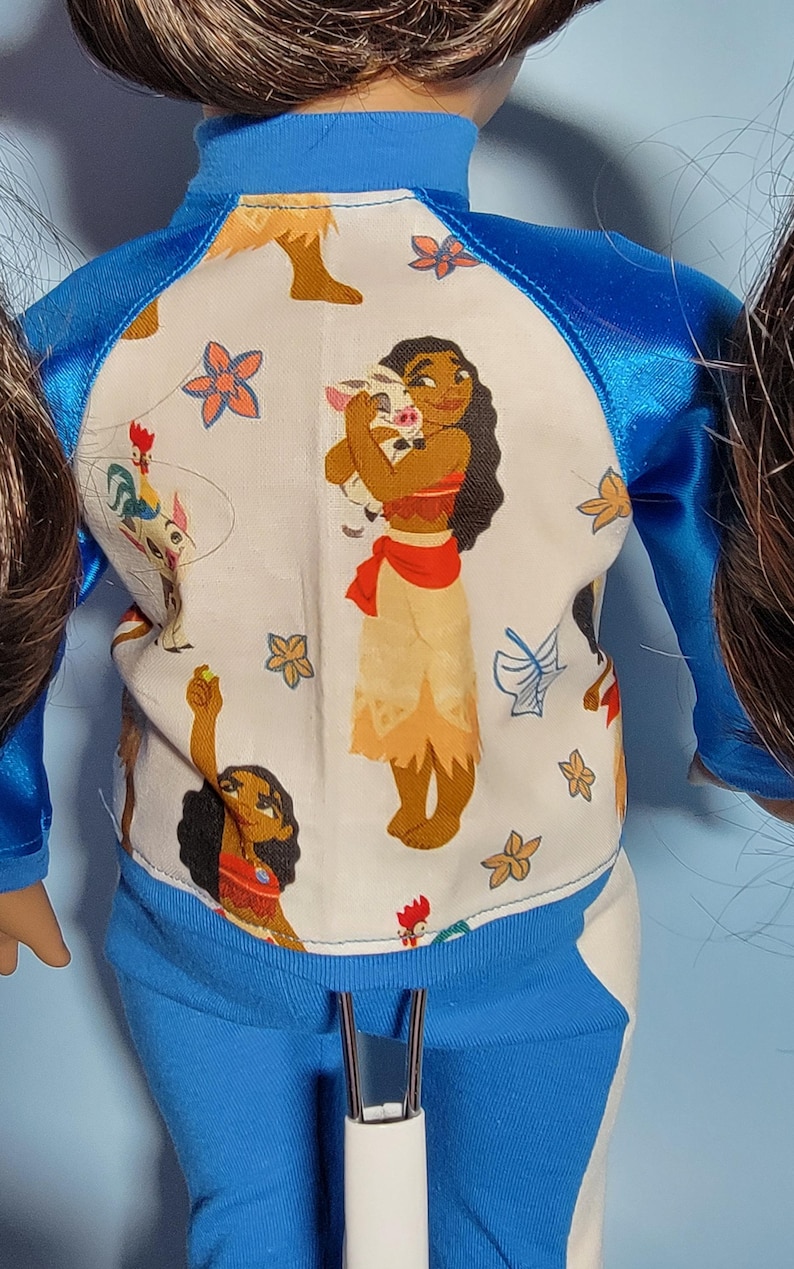 May include: A doll wearing a blue and white outfit with a character design. The top is white with a blue trim and features a cartoon illustration. The pants are solid blue. The outfit is designed for a doll.