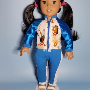 May include: A doll wearing a blue and white athletic outfit. The jacket has blue sleeves and a white body with a colorful print. The pants are blue with white stripes. The doll has dark hair in pigtails and is wearing white sandals.