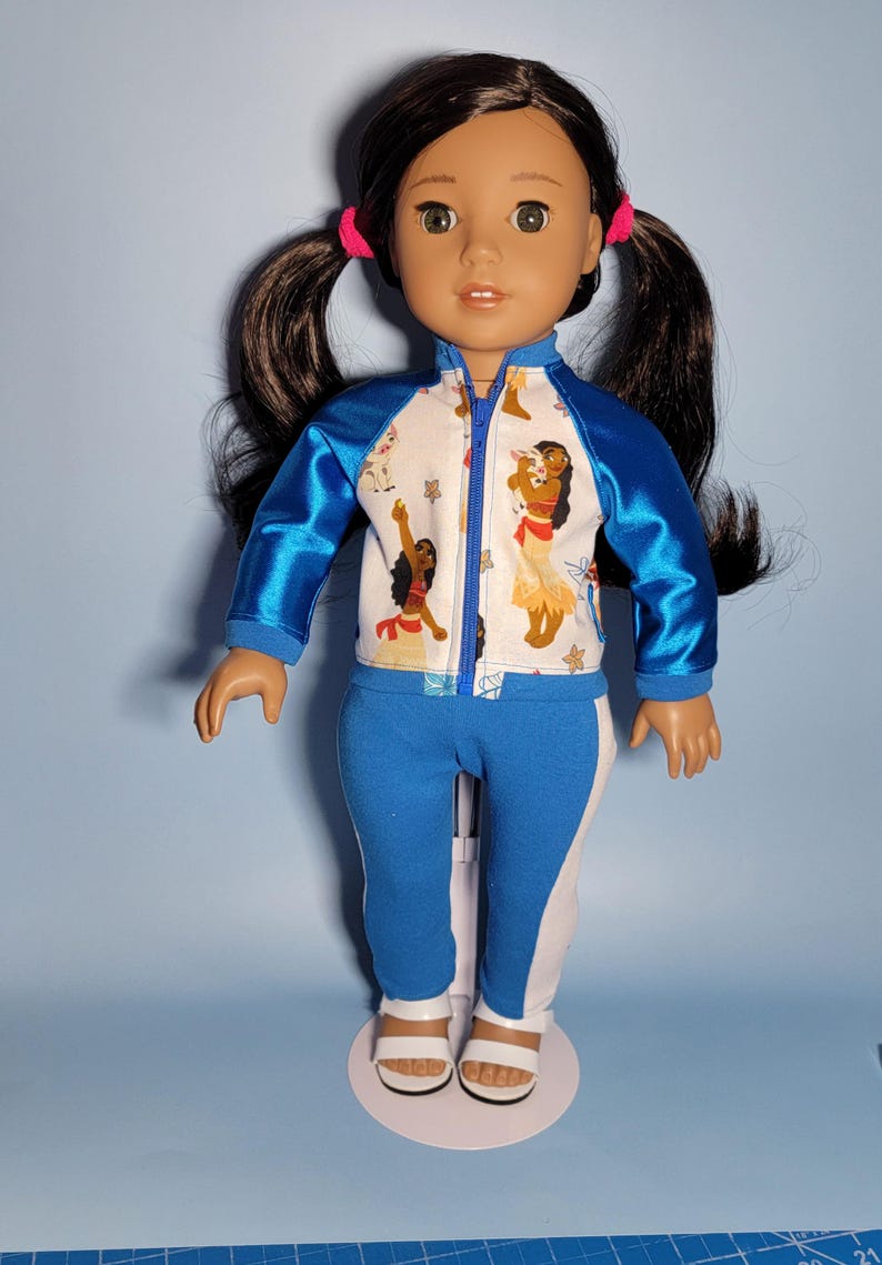 May include: A doll wearing a blue and white athletic outfit. The jacket has blue sleeves and a white body with a colorful print. The pants are blue with white stripes. The doll has dark hair in pigtails and is wearing white sandals.