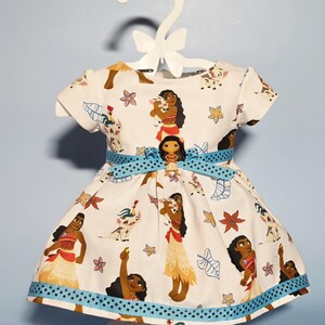 May include: A white dress with a blue ribbon belt and trim, featuring a repeating pattern of a cartoon character and floral designs. The dress is displayed on a white hanger.