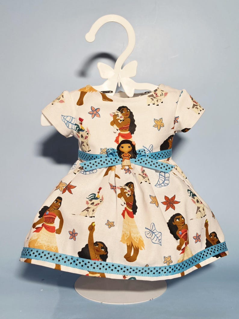 May include: A white dress with a blue ribbon belt and trim, featuring a repeating pattern of a cartoon character and floral designs. The dress is displayed on a white hanger.