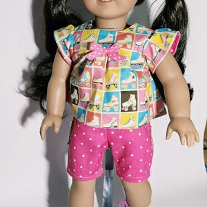 May include: A doll with dark hair in pigtails wearing a short-sleeved top featuring a grid pattern of colorful roller skates and a small pink bow. The doll is dressed in bright pink shorts with white polka dots and metallic purple roller skates.