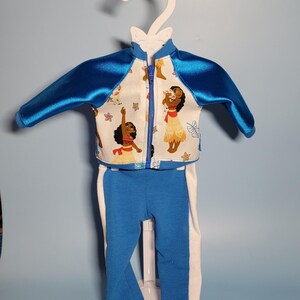 May include: A two-piece doll outfit featuring a blue and white color scheme. The jacket has blue satin sleeves and a white front with a cartoon character print. The pants are blue with white side stripes. The outfit is displayed on a white hanger.