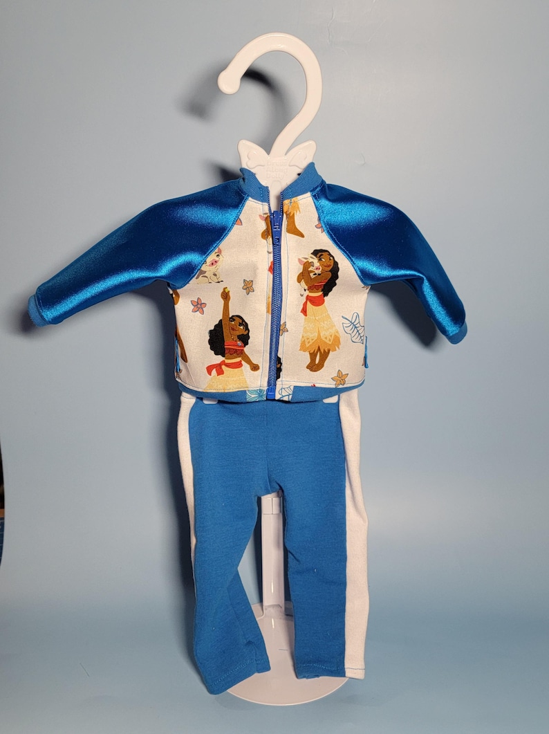 May include: A two-piece doll outfit featuring a blue and white color scheme. The jacket has blue satin sleeves and a white front with a cartoon character print. The pants are blue with white side stripes. The outfit is displayed on a white hanger.