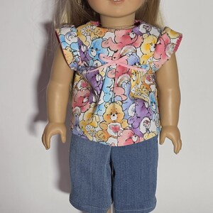 May include: A doll with blonde hair styled with two small clips, wearing a short-sleeved top featuring a colorful Care Bears print with a small pink bow at the neckline, paired with blue denim shorts and white slip-on shoes with gold star patterns.