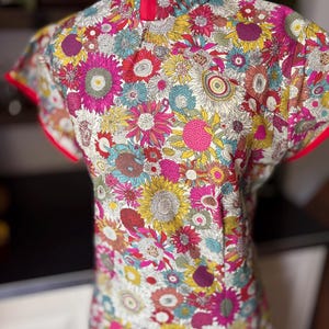 May include: A short-sleeved top with a mandarin collar featuring a vibrant, dense floral pattern in shades of pink, yellow, teal, and white. The garment has bright pink piping along the collar and sleeve edges, creating a distinct contrast.