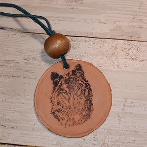 May include: A terracotta round pendant with a black and white wolf image, hanging from a teal cord with a brown wooden bead.