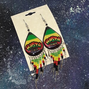 Peut inclure: Une paire de boucles d'oreilles pendantes avec un motif noir, vert, jaune et rouge. Les boucles d'oreilles présentent un charme en forme de larme avec les mots "Rasta Power" imprimés dessus. Les charmes sont attachés à une chaîne de perles avec des perles noires, vertes, jaunes et rouges.