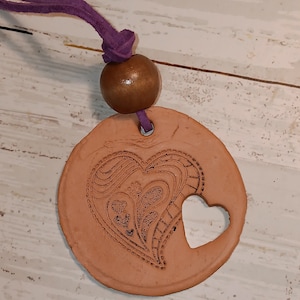 May include: A terracotta pendant necklace with a heart-shaped cutout and etched heart design. It has a purple suede cord, a wooden bead, and is set against a white-washed wooden surface. The pendant is round with a heart-shaped cutout.
