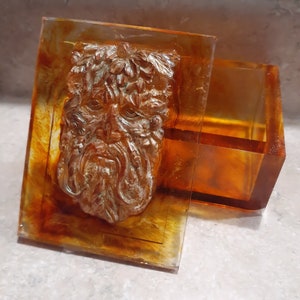 May include: A resin box with a green man face design. The box is clear with an amber tint and the face is detailed with leaves and a beard.