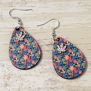 Peut inclure: Une paire de boucles d'oreilles pendantes avec une base en bois en forme de larme. La base est recouverte d'un motif coloré de feuilles de marijuana vertes, orange et bleues. Chaque boucle d'oreille présente un petit charme en argent en forme de feuille de marijuana avec des strass.