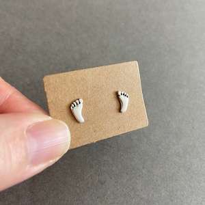 May include: A pair of silver stud earrings shaped like footprints. The earrings are displayed on a brown card.