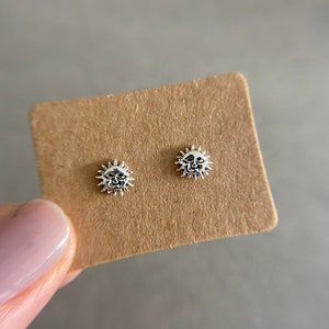 May include: A pair of silver earrings with a sun design. The earrings are on a brown card.