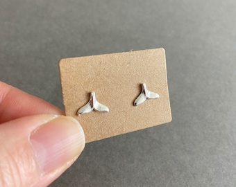 Pode incluir: Um par de brincos de prata com a forma de cauda de baleia em um cartão marrom.