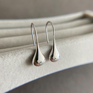 Könnte beinhalten: Ein Paar silberne Tropfenohrringe. Die Ohrringe haben eine polierte, reflektierende Oberfläche und ein einfaches Haken-Design. Die Tropfenform ist glatt und abgerundet und spiegelt die Umgebung wider. Die Ohrringe werden auf einer hellgrauen Oberfläche präsentiert.
