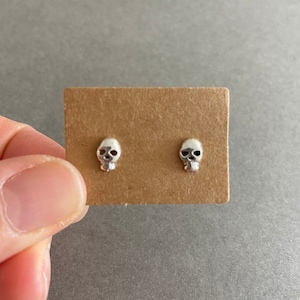 Op de afbeelding: Een paar kleine, zilverkleurige schedeloorbellen op een bruine kaart. De oorbellen hebben gedetailleerde oogkassen, neus en kaak. De achtergrond is effen grijs. De oorbellen zijn waarschijnlijk van metaal.