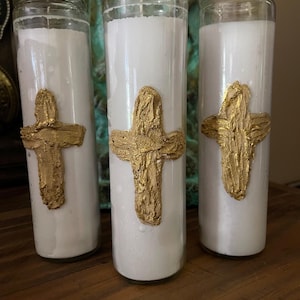 May include: Three white pillar candles in clear glass jars, each adorned with a textured gold cross. The candles are arranged on a wooden surface, with a blurred background.