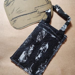 May include: A black and white feather print fabric tag with a matching handle is attached to a brown paper tag. The fabric tag is rectangular and has a raw edge. The paper tag is a light brown color.