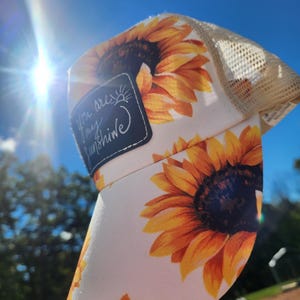May include: A white trucker hat with a sunflower print and a mesh back. The hat features a patch with the text "You are my sunshine". The sunflowers are yellow and orange, with a dark brown center.