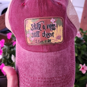 May include: A burgundy baseball cap with a mesh back. The front features a patch with the text "Still a mess, still chosen" and "1 Cor. 1:27" surrounded by sunflowers and flowers.