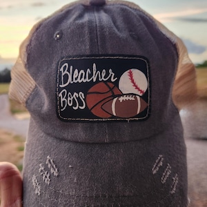 May include: Gray baseball cap with a mesh back and a distressed design. The cap features a patch with the words "Bleacher Boss" and illustrations of a baseball, basketball, and football.