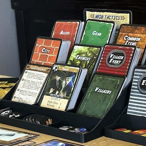 May include: A black tiered plastic card organiser holding various game cards labelled Mob Tactics, The Mob, City, Gear, Common Items, Enemies, Villain Event, Villains, Event, and Locations, with stacks of blue circular tokens and game boards nearby.
