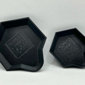 May include: Two black, pentagonal-shaped trays with raised edges. Each tray features a textured surface and the embossed text "BOARD GAME BOSS" centered on the base. The trays are identical in design but differ in their overall physical size.
