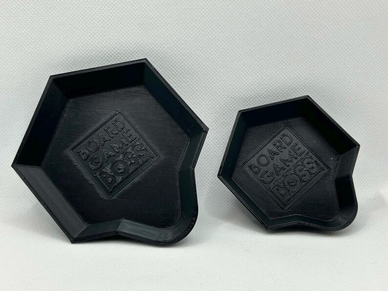 May include: Two black, pentagonal-shaped trays with raised edges. Each tray features a textured surface and the embossed text "BOARD GAME BOSS" centered on the base. The trays are identical in design but differ in their overall physical size.