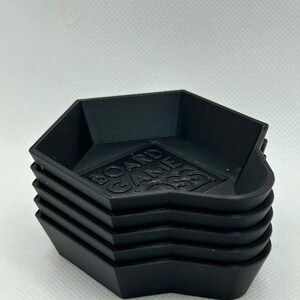 May include: A stack of five black hexagonal plastic trays with raised edges. The interior base of the top tray features the embossed text BOARD GAME BOSS in a block font. The trays have a matte finish and a layered texture from 3D printing.