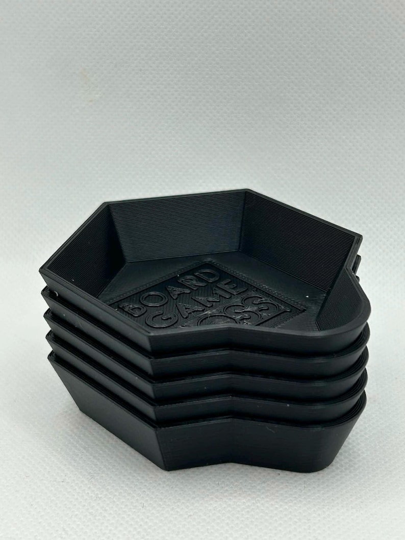 May include: A stack of five black hexagonal plastic trays with raised edges. The interior base of the top tray features the embossed text BOARD GAME BOSS in a block font. The trays have a matte finish and a layered texture from 3D printing.