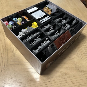 May include: A compartmentalized game organizer for the board game "Heroes of Barcadia." The organizer is filled with game pieces, including dice, cards, and miniature figures. The box is beige with black compartments and a brown leather-like accent.