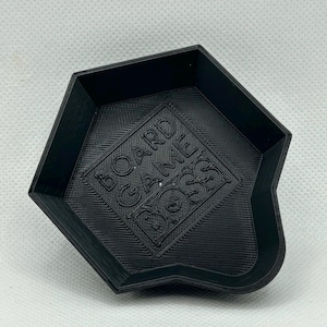 May include: A black, pentagonal-shaped plastic tray with raised edges and a tapered pouring spout. The interior base features the embossed text BOARD GAME BOSS centered within a square frame, displaying a textured, layered 3D printed finish.