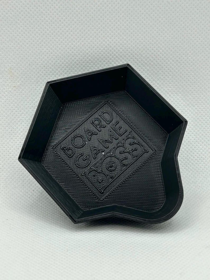 May include: A black, pentagonal-shaped plastic tray with raised edges and a tapered pouring spout. The interior base features the embossed text BOARD GAME BOSS centered within a square frame, displaying a textured, layered 3D printed finish.