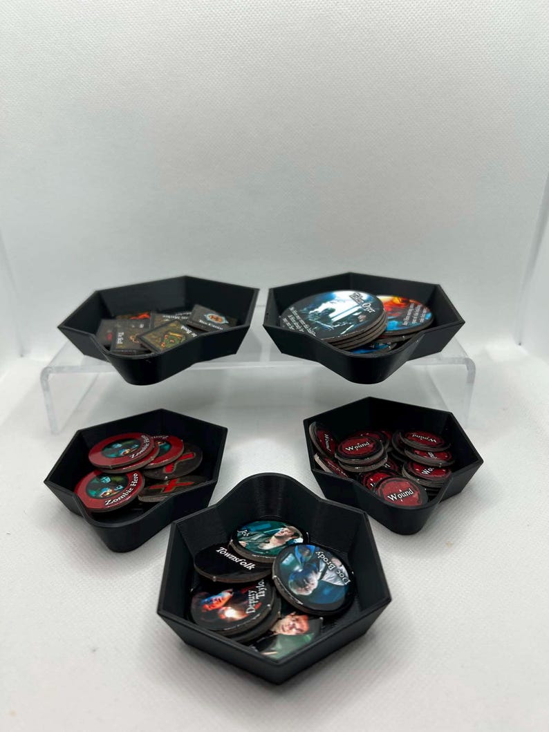 May include: Five black hexagonal plastic trays containing various circular and square game tokens. The tokens feature printed illustrations of characters, text labels such as Wound, Townfolk, and Zombie Here, and dark, colorful graphic designs.