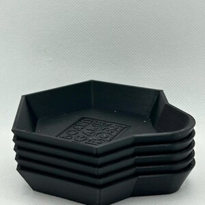 May include: A stack of four black, hexagonal-shaped trays with raised edges. Each tray features a flat base with the embossed text "BOARD GAME BOSS" centered on the bottom surface. The trays have a matte finish and a uniform, geometric design.