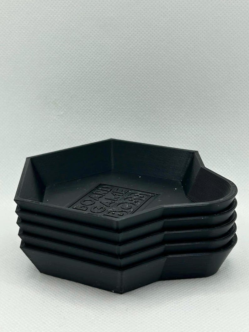 May include: A stack of four black, hexagonal-shaped trays with raised edges. Each tray features a flat base with the embossed text "BOARD GAME BOSS" centered on the bottom surface. The trays have a matte finish and a uniform, geometric design.