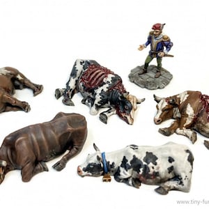 May include: A collection of six painted miniature cows, some with blood splatters, lying on a white surface. One of the cows has a bell around its neck. A miniature figure of a man in a red hat and blue and gold clothing stands on a stone base.